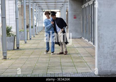 Zwei männliche Freunde gehen Arm in Arm auf dem Campus-Flur, tragen Mortarboard, Kleid und Hoodie Stockfoto