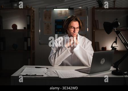 Fokussierter Arzt in Uniform, der am Tisch sitzt und in der Nachtschicht mit Laptop arbeitet. Kopierbereich Stockfoto