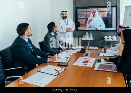 Verschiedene Geschäftsteams arbeiten an einer modernen Büro- und Videokonferenz mit einem arabischen Kollegen zusammen, der aus der Ferne beiträgt und die Teammitglieder des globalen Unternehmens präsentiert Stockfoto