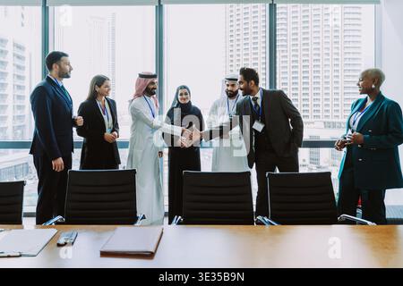 Vielfältige Geschäftsleute in einem modernen Büro in dubai, zwei Männer, die durch ein Skyline-Fenster die Hand schütteln, symbolisieren internationale Partnerschaft Stockfoto