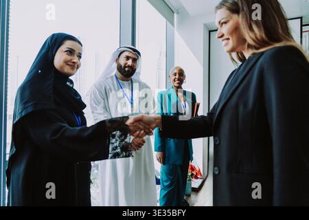 Zwei verschiedene Geschäftsfrauen schütteln in einem modernen Büro in dubai die Hand, um eine professionelle Partnerschaft zu festigen, während andere internationale Teammitglieder Zeugen davon sind Stockfoto