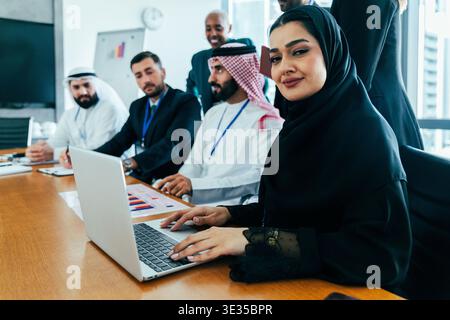 Arabische Frau, die einen Hijab trägt und lächelt, während sie auf einem Laptop schreibt, umgeben von multiethnischen Firmenkollegen, die an einem Geschäftstreffen in einem modernen o teilnehmen Stockfoto