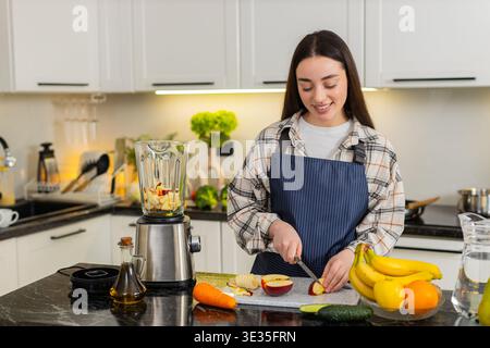 Junge Frau in Schürze schneidet Apfel in Scheiben und bereitet Zutaten für Smoothie zu, indem sie in der Nähe des Mixers zu Hause Obst hinzufügt. Gesundes Frühstück mit Bananen, Äpfeln und frischen Produkten auf der Küchenzeile. Stockfoto