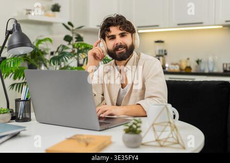 Der indische Mann zu Hause arbeitet an einem Laptop mit schnurlosen Kopfhörern und hört Podcast-Lächeln beim Tippen. Ein arabischer Typ, der am Tisch sitzt, tippt im Rhythmus der Musik die Tasten und bewegt die Schultern leicht und fühlt Freude und Konzentration. Stockfoto