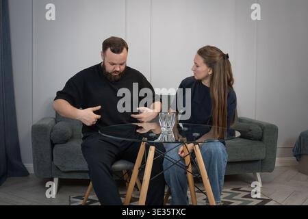 Ein bärtiger Mann erklärt der Frau Unbehagen, zeigt auf seinen Bauch, während das Paar in einem Wohnzimmer sitzt und sich in einem ernsten, aber zwanglosen Gespräch verwickelt Stockfoto