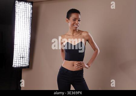 Eine lächelnde schwarze Frau mit kurzen Haaren posiert selbstbewusst in einem professionellen Fotostudio. Sie trägt ein schwarzes Korsett-Top und Jeans und lächelt an Stockfoto