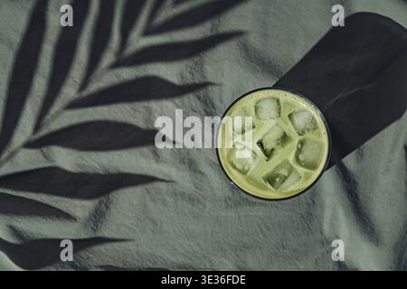 Sommertageszene mit Eismatcha Latte und tropischem Palmenblattschatten auf grünem Leinenhintergrund. Sonne und Schatten. Minimaler Sommermatcha. Stockfoto