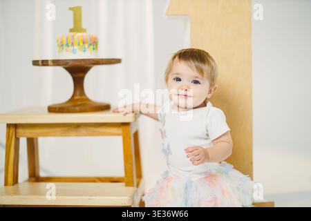 Ein Kind feiert seinen ersten Geburtstag. Das Kind trägt ein leichtes Outfit und steht neben einem Tisch mit einem Kuchen. Die Nummer eins ist displa Stockfoto