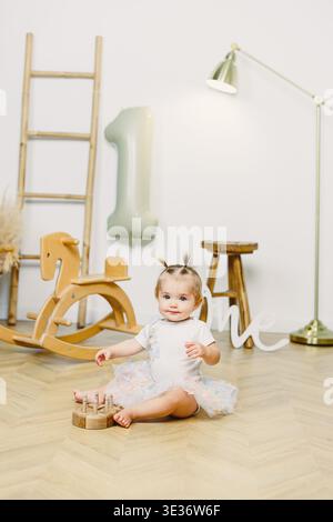 Ein Kind sitzt auf einem Holzboden neben einem Schaukelpferd. Das Kind spielt mit einem Spielzeug, während ein großer Ballon die Nummer eins im Hintergrund zeigt. Die s Stockfoto