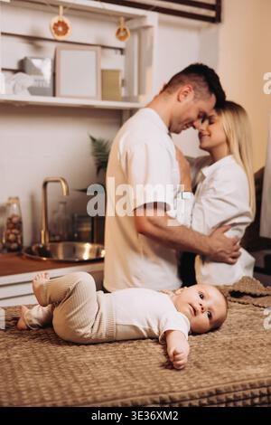 In einer gemütlichen Küche teilt ein liebevolles Paar einen süßen Moment, während ihr liebenswertes Baby in der Nähe spielt und die Familienliebe und die Freude der Elternschaft in Ever verkörpert Stockfoto