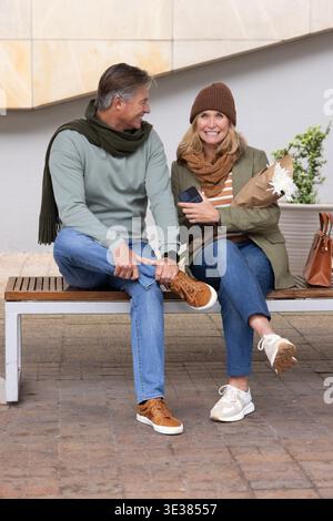 Seniorenpaar, das auf einer Holzbank auf dem städtischen plaza sitzt und einen Blumenstrauß und ein Smartphone hält Stockfoto