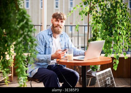 Man arbeitet mit Laptop und Smartphone am Tisch im Freien, mit einem Glas Eiskaffee. Solarpanel in der Nähe, der nachhaltigen, modernen, umweltfreundlichen Arbeitsbereich betont, der Technologie und Nachhaltigkeit vereint. Stockfoto