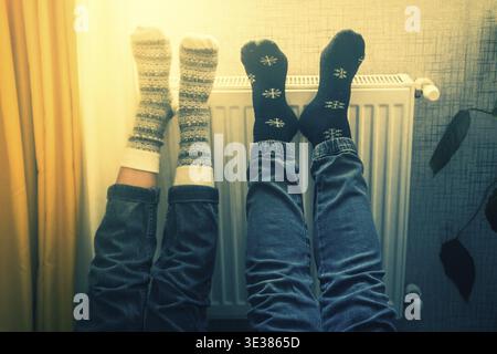 Ein Paar hält die Beine hoch und heizt die Füße in kalten Wohnungen drinnen auf der Heizung im Winter mit kuscheligen, stilvollen Wollsocken an. Valentinstag lustig zusammen warm f Stockfoto