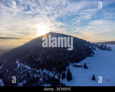 Der Berg Bukovec liegt im Winter über dem Dorf Jizerka im Isergebirge. Schnee bedeckt den Boden und die Bäume. Die Sonne untergeht im Hintergrund und verleiht der Szene Licht. Stockfoto
