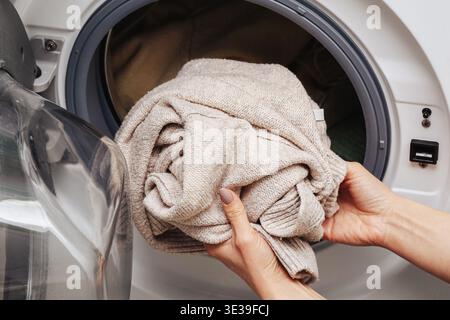 Frau, die einen beigefarbenen Pullover in die Waschmaschine steckt. Wäschereiroutine zu Hause. Haushaltsarbeiten im modernen Badezimmer zu Hause. Stockfoto