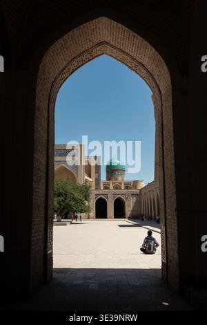 Blick durch ein gewölbtes Tor auf die Kalyan Complex Madrasa mit ihrer türkisfarbenen Kuppel und dem sonnendurchfluteten Innenhof unter klarem blauem Himmel. Stockfoto