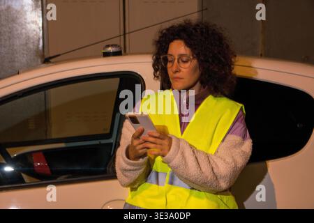 Frau in Warnweste und Brille, die auf dem Smartphone neben einem kaputten Auto nachts in einer urbanen Umgebung um Pannenhilfe bittet Stockfoto