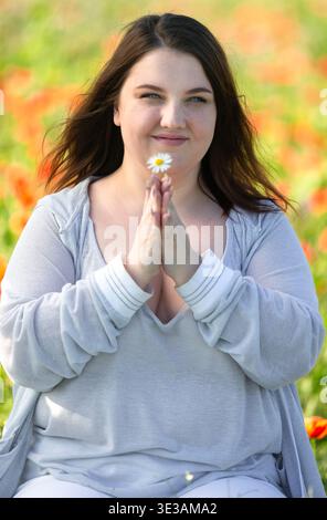 Porträt einer jungen Frau, die den Duft einer Wildblume einatmet. Hände gefaltet im Gebet. Vertikales Foto. Körperpositivität Stockfoto