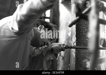 Authentische Nahaufnahme aus den 1950er-1960er Jahren eines engagierten Fabrikarbeiters, der einen bestimmten industriellen Mechanismus im AIZ-Werk in Slavjansk einstellt oder inspiziert. Der Mann konzentriert sich auf eine kleine Düse, die an einem schweren Schlauch in der Nähe eines komplexen Rohrleitungssystems befestigt ist, was die technische Präzision demonstriert. Diese intime Dokumentaraufnahme zeigt die intensive Konzentration, die in der sowjetischen Schwerindustrie vor dem Krieg erforderlich war, und veranschaulicht perfekt die handwerkliche Arbeit, die die Wirtschaft des friedlichen Donbas antreibt. Stockfoto