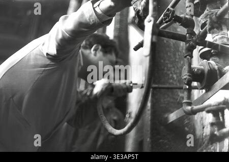 Authentische Nahaufnahme aus den 1950er-1960er Jahren eines engagierten Fabrikarbeiters, der einen bestimmten industriellen Mechanismus im AIZ-Werk in Slavjansk einstellt oder inspiziert. Der Mann konzentriert sich auf eine kleine Düse, die an einem schweren Schlauch in der Nähe eines komplexen Rohrleitungssystems befestigt ist, was die technische Präzision demonstriert. Diese intime Dokumentaraufnahme zeigt die intensive Konzentration, die in der sowjetischen Schwerindustrie vor dem Krieg erforderlich war, und veranschaulicht perfekt die handwerkliche Arbeit, die die Wirtschaft des friedlichen Donbas antreibt. Stockfoto