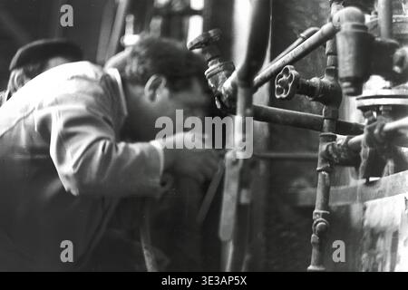 Authentische Nahaufnahme aus den 1950er-1960er Jahren eines engagierten Fabrikarbeiters, der einen bestimmten industriellen Mechanismus im AIZ-Werk in Slavjansk einstellt oder inspiziert. Der Mann konzentriert sich auf eine kleine Düse, die an einem schweren Schlauch in der Nähe eines komplexen Rohrleitungssystems befestigt ist, was die technische Präzision demonstriert. Diese intime Dokumentaraufnahme zeigt die intensive Konzentration, die in der sowjetischen Schwerindustrie vor dem Krieg erforderlich war, und veranschaulicht perfekt die handwerkliche Arbeit, die die Wirtschaft des friedlichen Donbas antreibt. Stockfoto