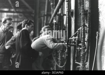 Authentische Nahaufnahme aus den 1950er-1960er Jahren eines engagierten Fabrikarbeiters, der einen bestimmten industriellen Mechanismus im AIZ-Werk in Slavjansk einstellt oder inspiziert. Der Mann konzentriert sich auf eine kleine Düse, die an einem schweren Schlauch in der Nähe eines komplexen Rohrleitungssystems befestigt ist, was die technische Präzision demonstriert. Diese intime Dokumentaraufnahme zeigt die intensive Konzentration, die in der sowjetischen Schwerindustrie vor dem Krieg erforderlich war, und veranschaulicht perfekt die handwerkliche Arbeit, die die Wirtschaft des friedlichen Donbas antreibt. Stockfoto
