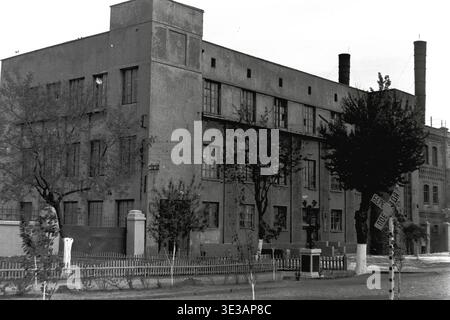 Vintage-Schwarzweiß-Archivfoto aus den 1950er oder 1960er Jahren mit einem riesigen mehrstöckigen Industriegebäude in der Armature and Insulator Plant in Slavjansk. Die historische Ziegelfassade hat zahlreiche große Fenster, während in der Nähe der asphaltierten Straße ein Bahnübergangsschild steht, das vor Zügen warnt. Weiß getünchte Bäume säumen den traditionellen Holzzaun. Ein authentischer Einblick in die Schwerindustrie, die sowjetische Fertigungsarchitektur, die Fabrikinfrastruktur und den Alltag im friedlichen Donbass. Stockfoto