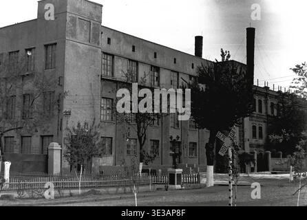 Vintage-Schwarzweiß-Archivfoto aus den 1950er oder 1960er Jahren mit einem riesigen mehrstöckigen Industriegebäude in der Armature and Insulator Plant in Slavjansk. Die historische Ziegelfassade hat zahlreiche große Fenster, während in der Nähe der asphaltierten Straße ein Bahnübergangsschild steht, das vor Zügen warnt. Weiß getünchte Bäume säumen den traditionellen Holzzaun. Ein authentischer Einblick in die Schwerindustrie, die sowjetische Fertigungsarchitektur, die Fabrikinfrastruktur und den Alltag im friedlichen Donbass. Stockfoto