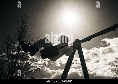 Monochrome Silhouette eines Kindes, das hoch gegen hellen Himmel mit warmem Sonnenlicht schwingt. Emotionale Szene, die Freiheit, Freude und sorglose Kindheit symbolisiert. Stockfoto