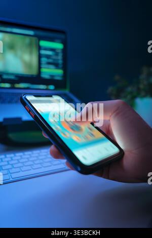 Nahaufnahme des Smartphones in der Hand und des schnellen Scrollens von Social-Media-Feeds in dunklen Räumen mit Schreibtisch auf Hintergrund, beleuchtet von blauem Licht. Digitale sucht und unsinniger Content-Konsum Stockfoto