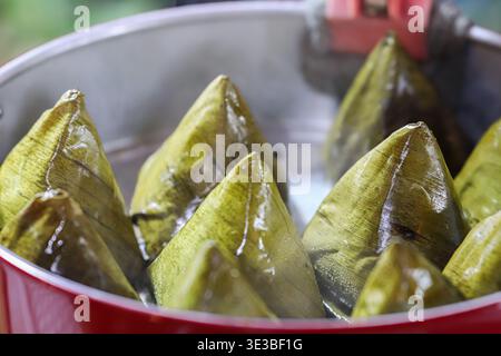 Traditionelle thailändische und chinesische Reisknödel, die in einem Metalltopf dampft. Heiße Dämpfe und Wassertropfen auf Bananenblättern während des Dragon Boat Festival. Stockfoto