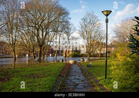 Ein Pfad führt durch einen Stadtpark zu einem überfluteten Gebiet durch einen Fluss mit Häusern im Hintergrund, City of York, England. Stockfoto