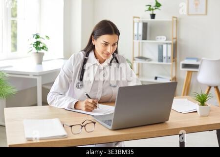 Lächelnde junge Ärztin in Uniform, die am Schreibtisch sitzt und mit einem Laptop in der Arztpraxis arbeitet. Stockfoto