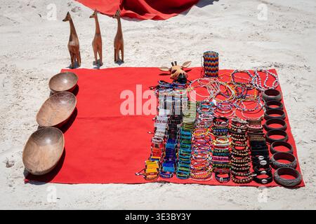 Traditioneller afrikanischer handgemachter Schmuck, perlenbesetzte Armbänder, Holzschalen und Giraffen-Figuren auf einem roten Tuch an einem weißen Sandstrand in Sansibar Stockfoto