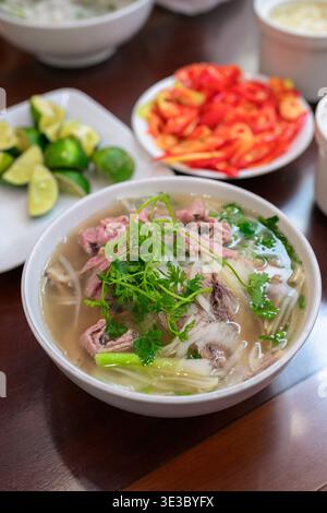 Eine Schüssel mit traditioneller vietnamesischer Rindfleischnudelsuppe, bekannt als Pho bo, serviert mit frischem Koriander und grünen Zwiebeln. Das Gericht wird von einer Beilage begleitet Stockfoto