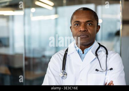 Erfahrener Arzt, der einen weißen Labormantel und ein Stethoskop über den Schultern trägt, mit überkreuzten Armen steht und Selbstvertrauen und Fachwissen in einer medizinischen Umgebung darstellt Stockfoto