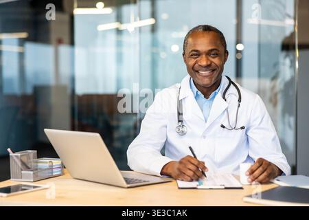 Reifer Arzt im Labormantel und Stethoskop, der selbstbewusst lächelt, während er an einem Schreibtisch mit Laptop und Dokumenten arbeitet und Professionalität und Expertise im Gesundheitswesen verkörpert Stockfoto