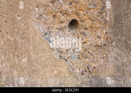 Nahaufnahme der verwitterten Betonoberfläche mit freiliegendem Zuschlagstoff und einem kleinen gebohrten Loch, das raue Textur, Verschleiß und industrielle Details zeigt. Stockfoto