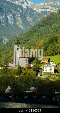 Eine malerische weiße Kirche mit einem Uhrenturm, eingebettet zwischen Herbstbäumen und traditionellen Häusern vor massiven Bergen. Stockfoto