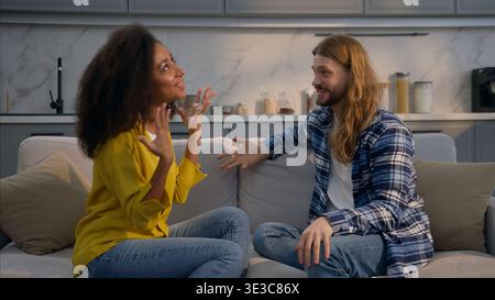 Glückliches aufgeregtes multiethnisches Paar erstaunliche Nachrichten zu Hause sprechende Couch Kaukasischer Mann überrascht hören Gespräch Afroamerikanerin lächelndes Wunder Stockfoto