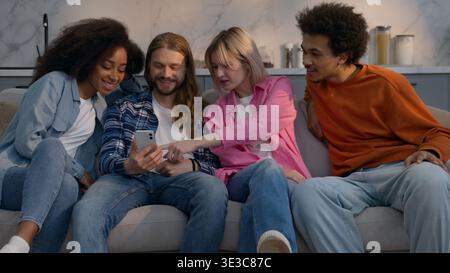 Kaukasischer Mann zeigt Handy-Video zu multiethnischen Freunden auf der Heimcouch lacht, spricht zusammen afroamerikanische Frauen Männer Stockfoto