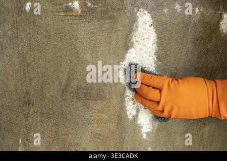 Nahaufnahme einer Person mit orangefarbenen Gummischutzhandschuhen, die eine schmutzige Betonwandoberfläche mit einem Stahlwolle-Pad, weißem Reinigungspulver-Schaum schrubbt Stockfoto