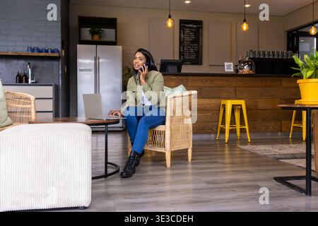 Indische Frau in Olivenjacke, die im Korbsessel im Café sitzt und Telefon hält und Laptop benutzt Stockfoto