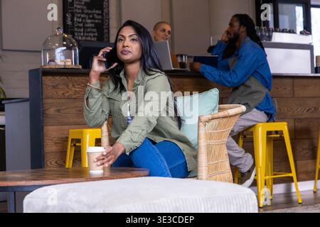 Indische Frau, die auf dem Smartphone spricht und nach einer Kaffeetasse zum Mitnehmen im Korbsessel im Café greift Stockfoto