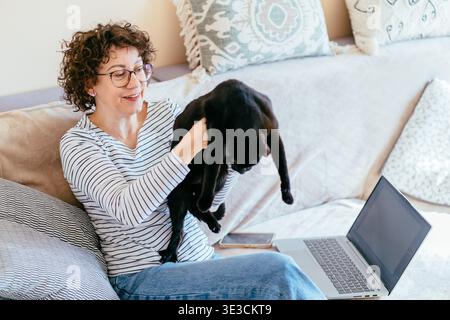 Frau mittleren Alters, die eine schwarze Katze hält, während sie zu Hause am Laptop arbeitet Stockfoto
