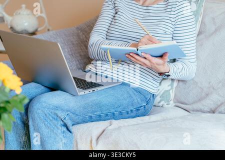 Frau mittleren Alters schreibt Notizen mit Laptop auf Sofa im Innenraum Stockfoto