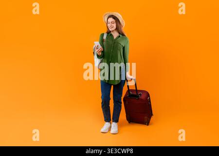 Junge Frau steht mit Koffer und Telefon bereit für die Reise in leuchtendem orangefarbenem Hintergrund Stockfoto