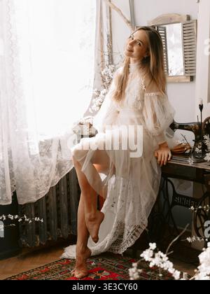 Wunderschöne Braut in Vintage-Spitzenkleid in der Nähe des Fensters, sanftes Licht romantisches Lifestyle-Porträt drinnen Stockfoto