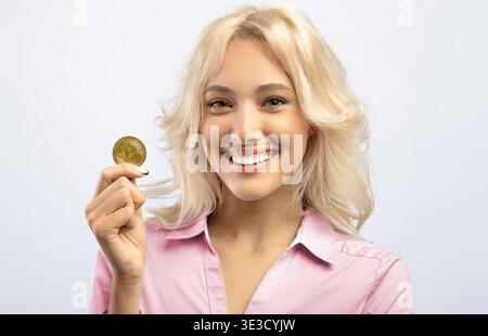 Eine Frau, die eine Goldmünze hält und während eines Studiodrehs in natürlichem Licht lächelt Stockfoto