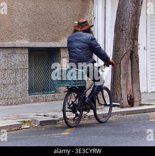 Ein Mann mit braunem Hut und grauer Jacke fährt mit einem Korb auf der Straße neben einer Steinmauer und einem Baum in einer städtischen Umgebung Stockfoto